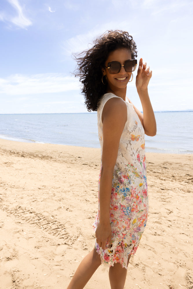 Side view of a woman in a white floral lace dress with colourful embroidered flowers, styled with sunglasses and sandals on a beach shoreline.