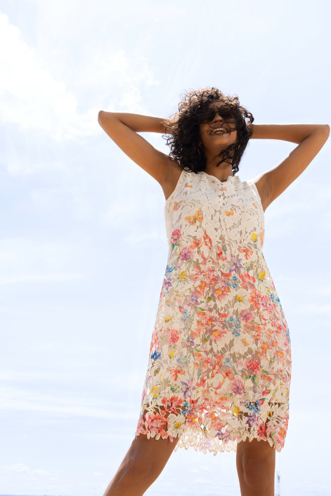 Low-angle view of a sleeveless white lace dress with vibrant floral embroidery, featuring a sheer overlay and scalloped hem detail.