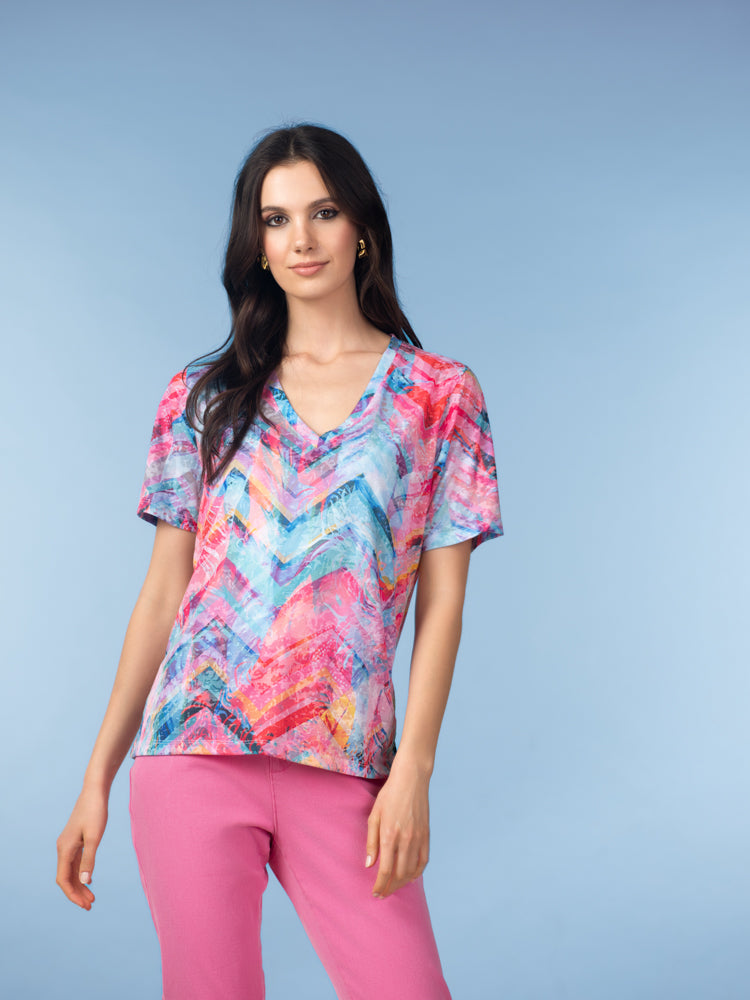 Woman modelling a short-sleeve V-neck T-shirt with an abstract chevron print in shades of pink, blue, and coral, paired with pink pants, against a soft blue studio background.