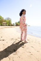 Model standing on a sandy beach wearing a pink and coral floral sheer blouse with matching wide-leg pants and white slide sandals, photographed from the back with the shoreline in the background.