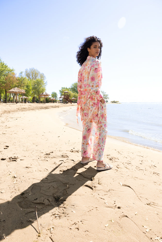 Model standing on a sandy beach wearing a pink and coral floral sheer blouse with matching wide-leg pants and white slide sandals, photographed from the back with the shoreline in the background.