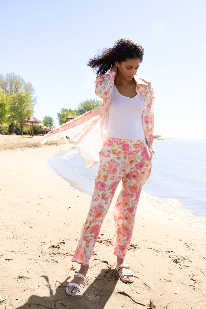 Model walking along the beach wearing a pink floral sheer button-front blouse over a white tank top, paired with matching pink floral straight-leg pants and white slide sandals.