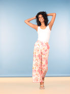 Woman in white tank top and pink floral wide-leg pants posing against a blue background.