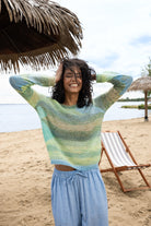 Woman standing on a sandy beach under a thatched umbrella, smiling with hands in her hair, wearing a soft green and blue striped lightweight knit sweater and loose light blue drawstring pants, with water and a lounge chair in the background.