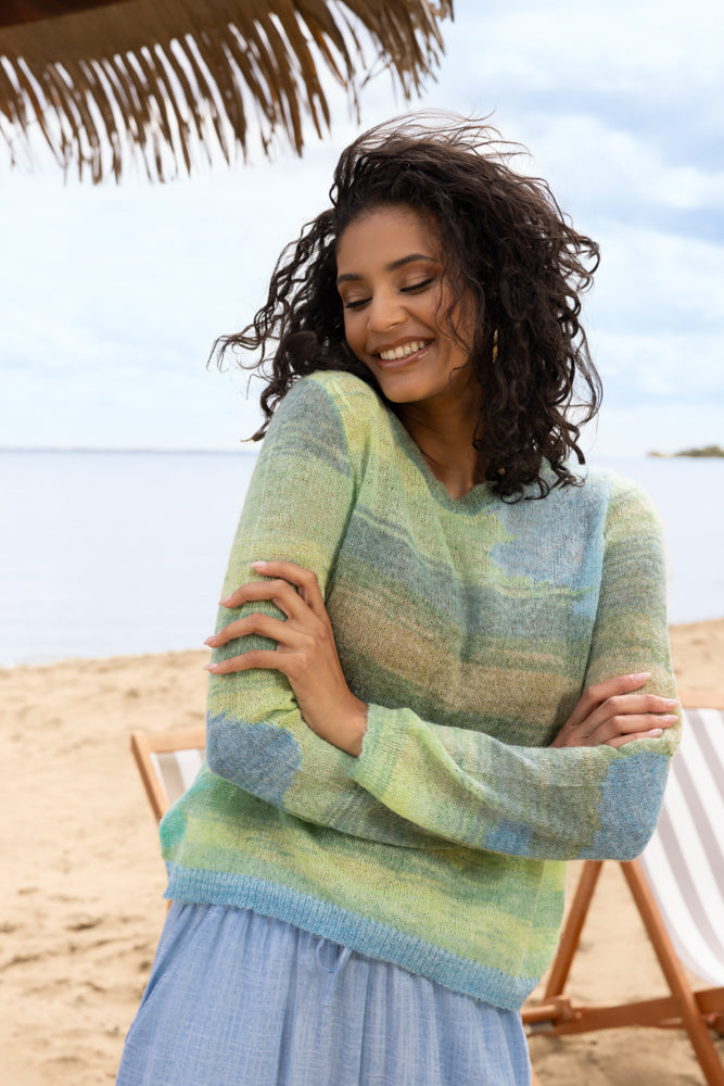 Close-up of a woman on the beach wearing a green and blue variegated knit sweater with long sleeves, styled with light blue flowy pants, smiling with arms gently crossed and water in the background.