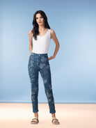 Woman wearing high-rise reversible slim jeans with a floral pattern, styled with a white sleeveless tank top and flat sandals, standing against a soft blue studio backdrop.