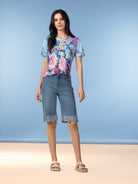 Model wearing light wash denim knee-length shorts with scalloped lace trim, paired with a floral short-sleeve top and flat sandals.