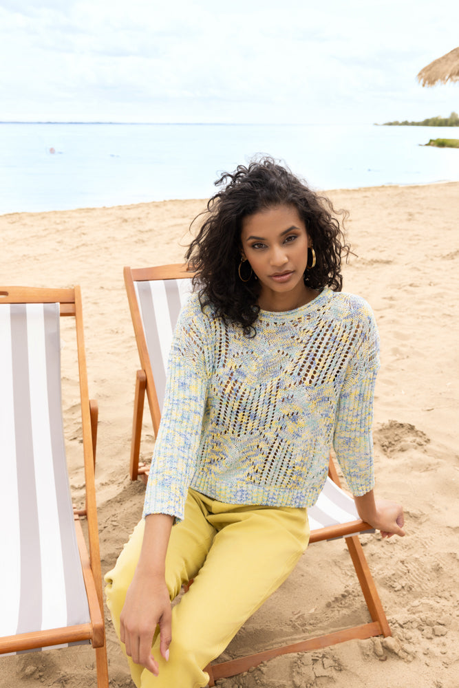Model wearing a blue and yellow melange open-knit sweater with three-quarter sleeves and crew neckline, styled with yellow trousers, seated on a beach deck chair.
