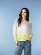Woman wearing a lightweight button-front cardigan in a soft ombré fade from ivory to chartreuse, styled with blue jeans against a light blue studio background.