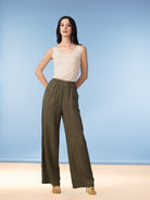 Full-length studio image of a woman wearing olive green wide-leg pants with a relaxed drape and elastic waistband, paired with a sleeveless beige tank top and strappy heels, posed against a soft blue backdrop.
