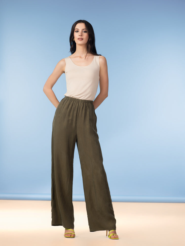 Full-length studio image of a woman wearing olive green wide-leg pants with a relaxed drape and elastic waistband, paired with a sleeveless beige tank top and strappy heels, posed against a soft blue backdrop.