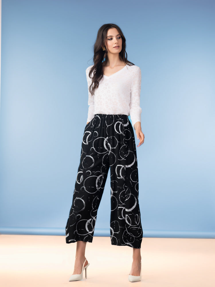 Model wearing a lightweight white V-neck sweater with long sleeves, styled with black wide-leg cropped pants featuring a circular pattern and paired with pointed heels, photographed against a soft blue studio background.