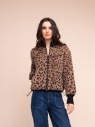 A woman modeling a cropped leopard-print jacket with a black zipper, black cuffs, and black drawstrings, paired with blue jeans against a solid beige background.