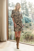 Woman wearing a zebra print dress and brown boots standing in a room with large windows.