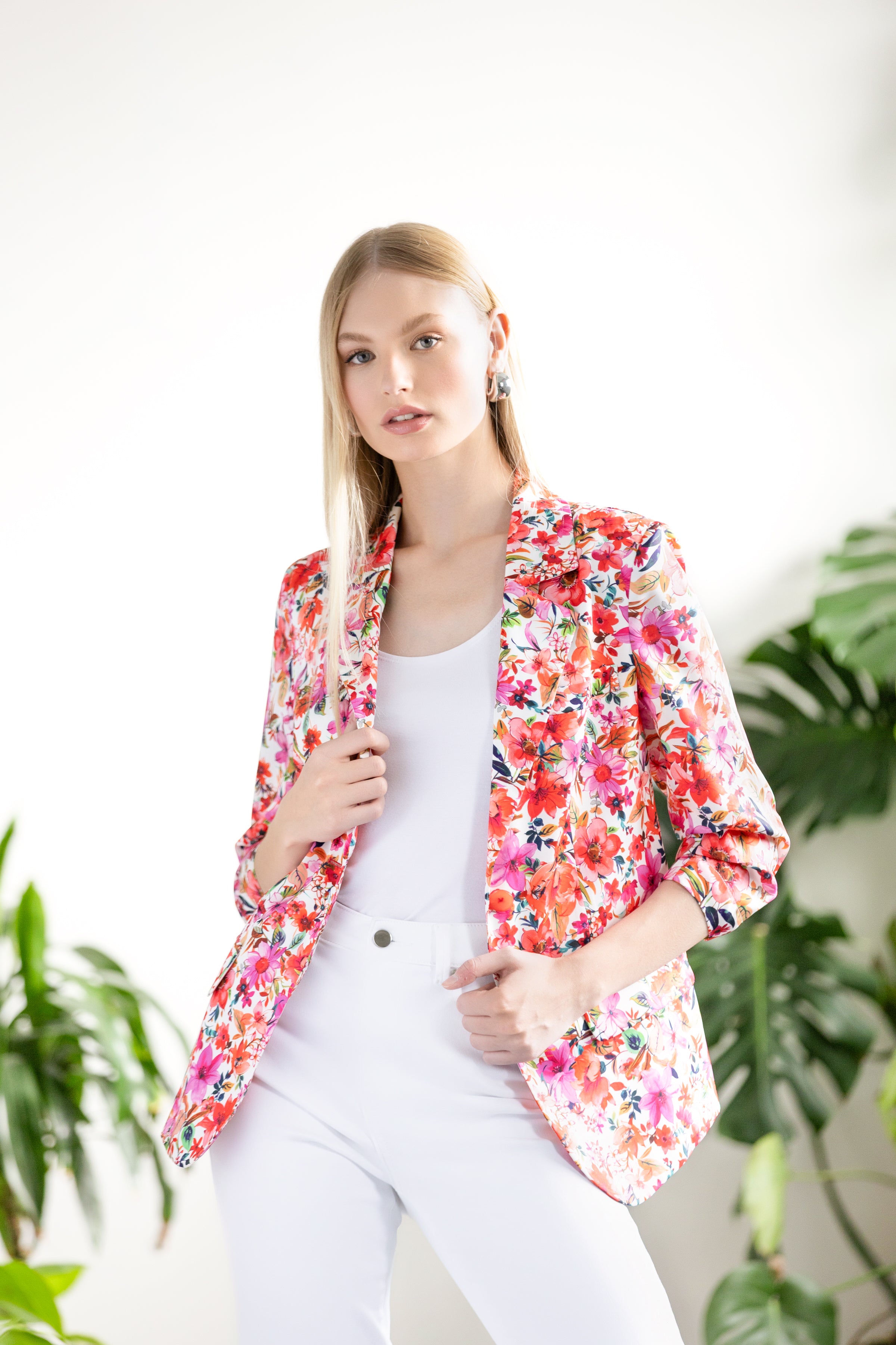 Woman wearing a floral blazer over a fitted top and high-waisted pants, standing in a bright studio with leafy plants iin the background.