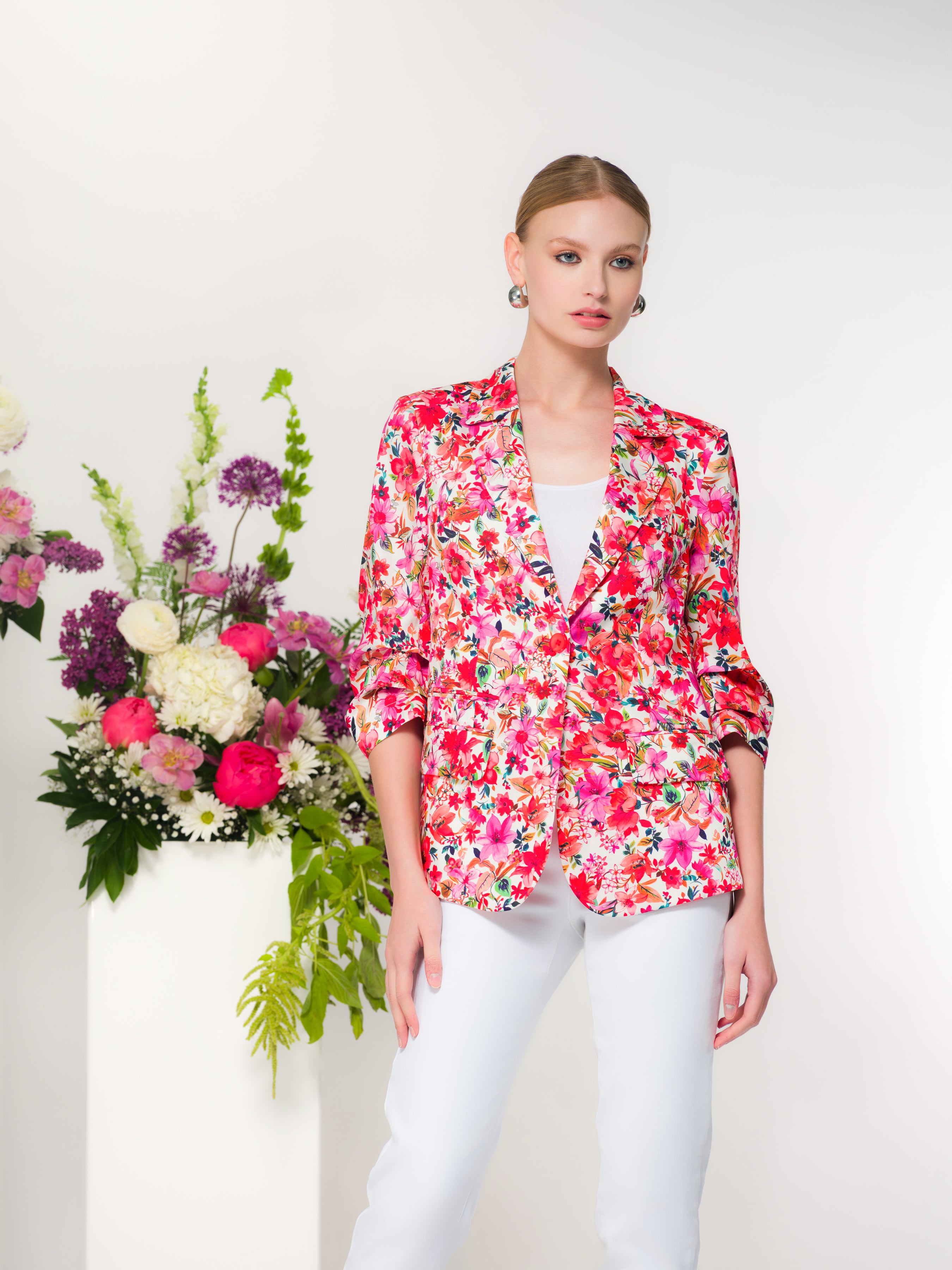 Woman wearing a floral blazer over a fitted top and high-waisted pants, standing next to a floral arrangement.