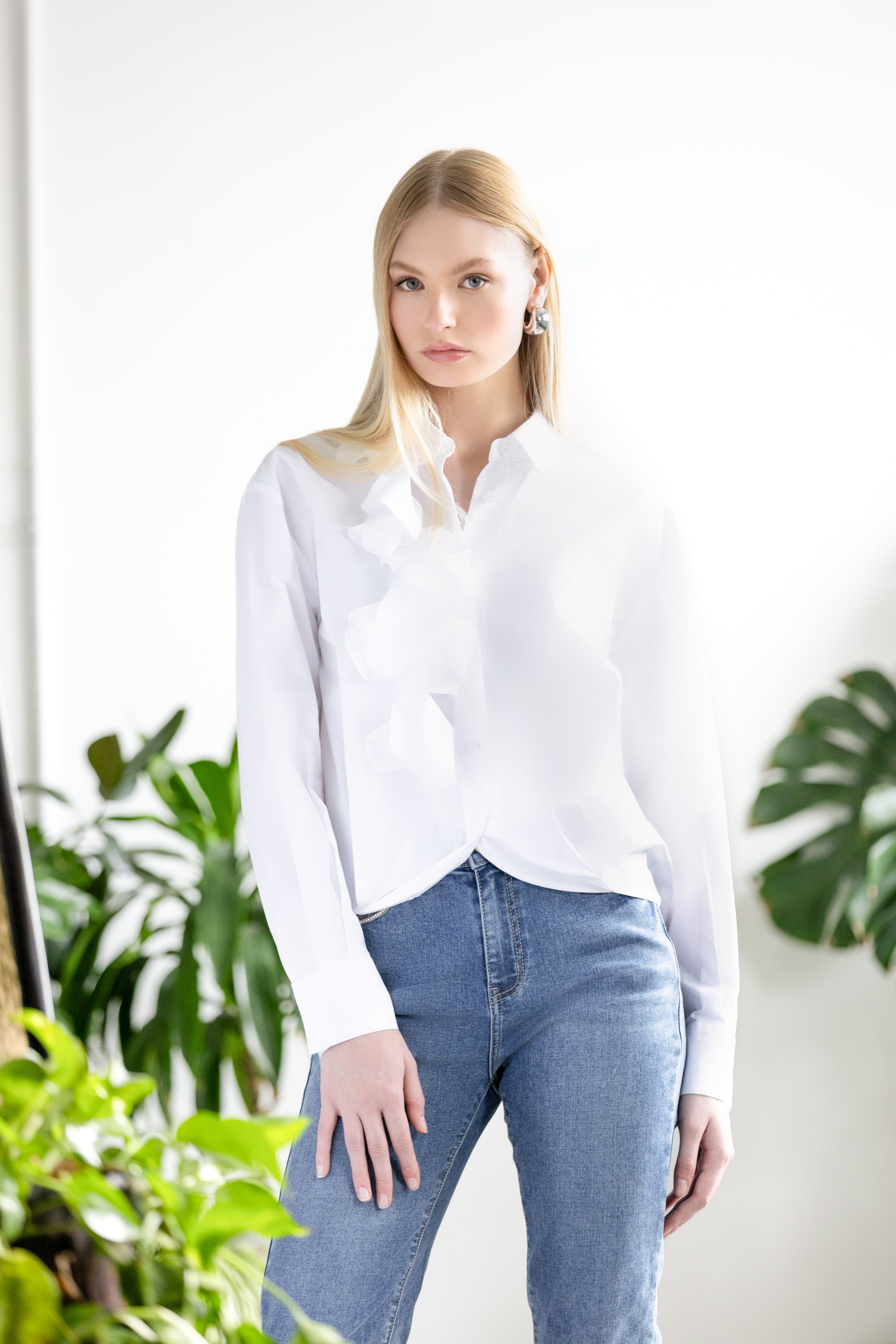 Woman with light hair wearing a white blouse and light jeans in a white studio surrounded by green leafy plants. 