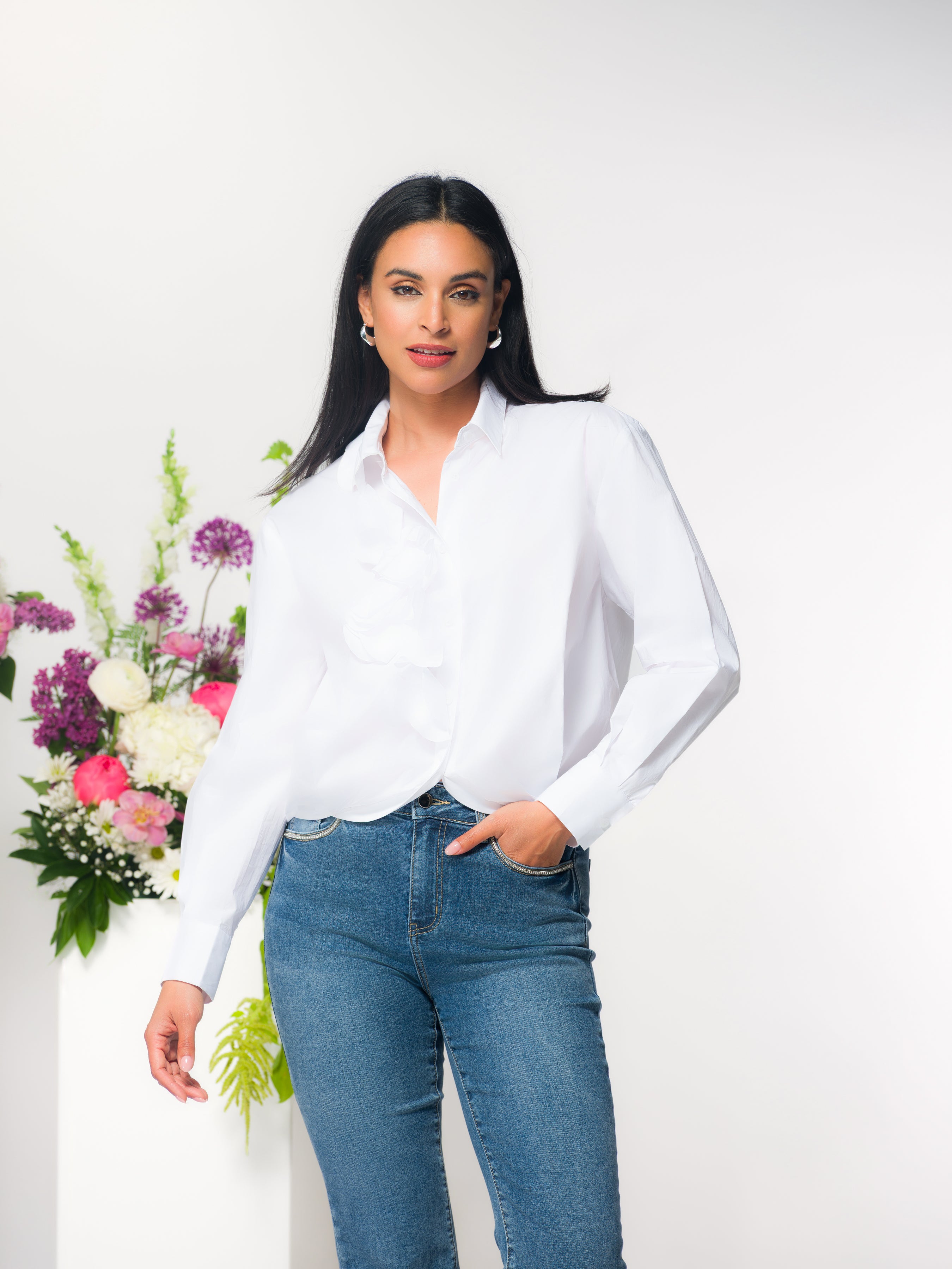 Woman with dark hair wearing a white blouse and denim jeans in a studio standing next to a floral arrangement.