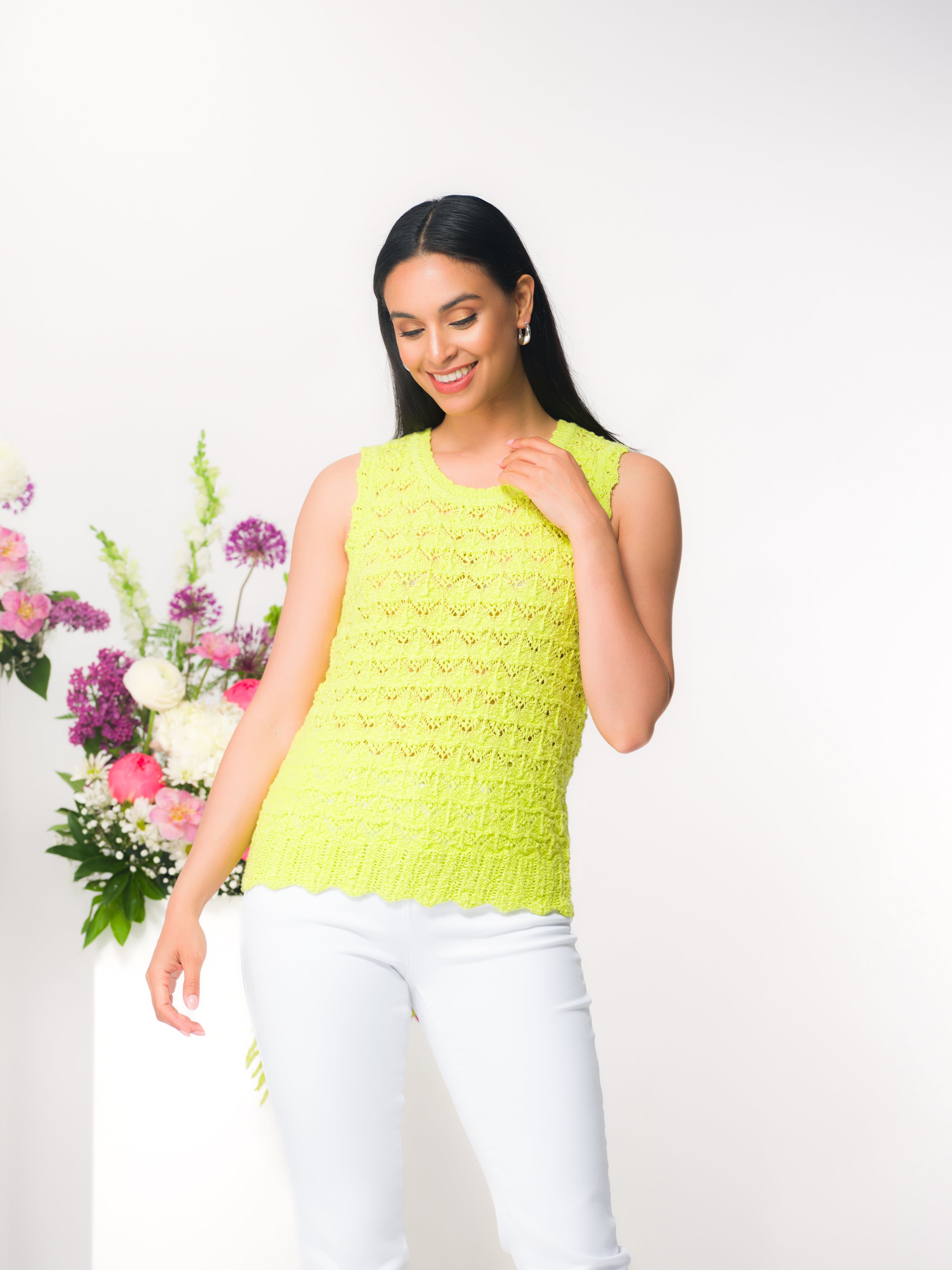 Woman wearing a sleeveless textured  lime knit top with a crew neckline and scalloped hem, paired with fitted pants, standing in a bright studio setting with floral accents in the background.