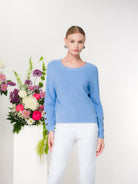 Woman wearing a blue sweater with white pants standing next to a floral arrangement with a white background