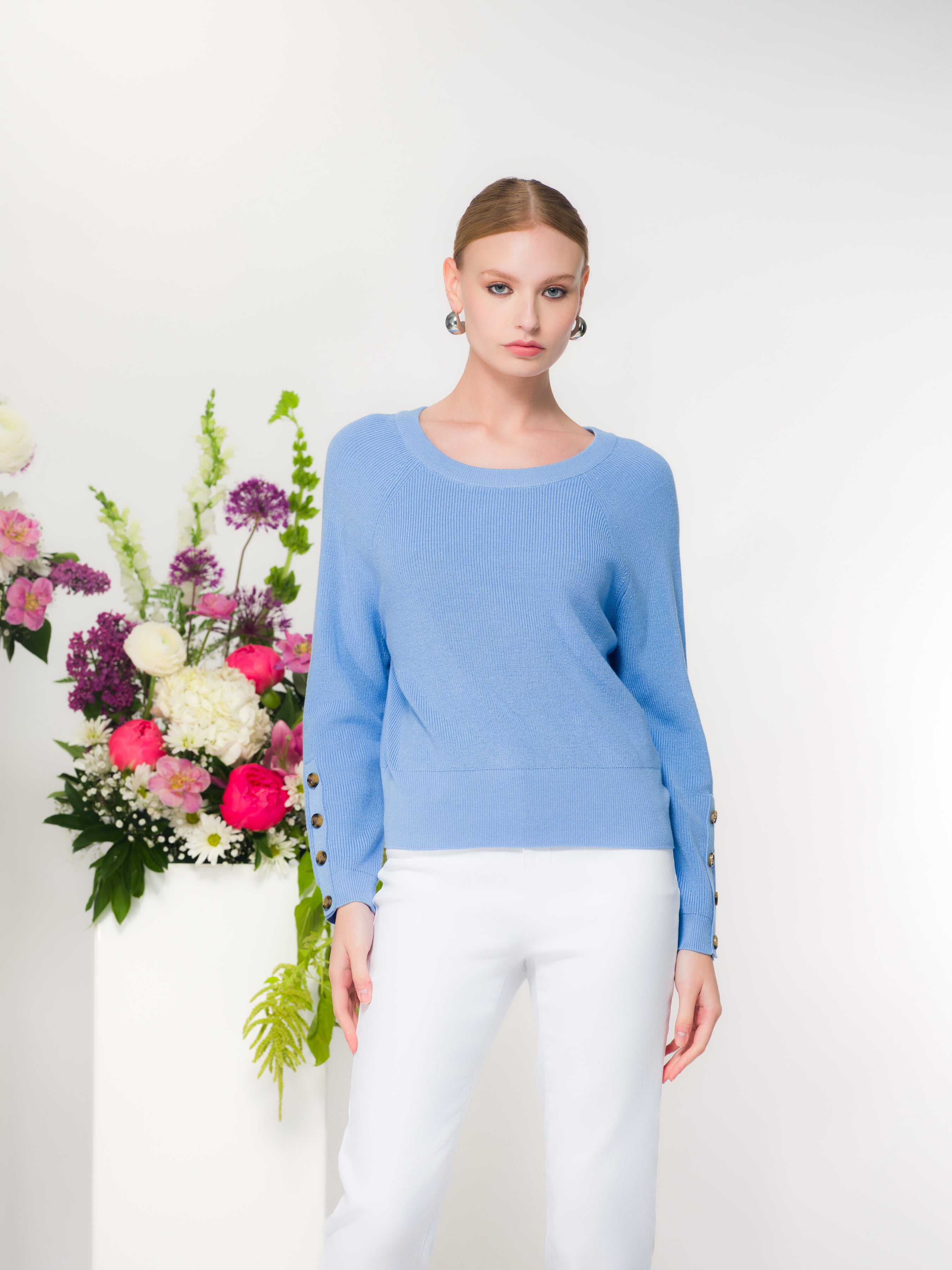 Woman wearing a blue sweater with white pants standing next to a floral arrangement with a white background