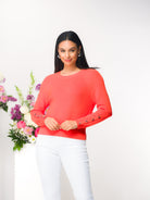 Woman wearing a coral sweater with white pants standing next to a floral arrangement with a white background