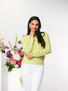 Woman wearing a lime green sweater with white pants standing next to a floral arrangement with a white background