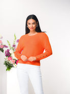 Woman wearing an orange sweater with white pants standing next to a floral arrangement with a white background