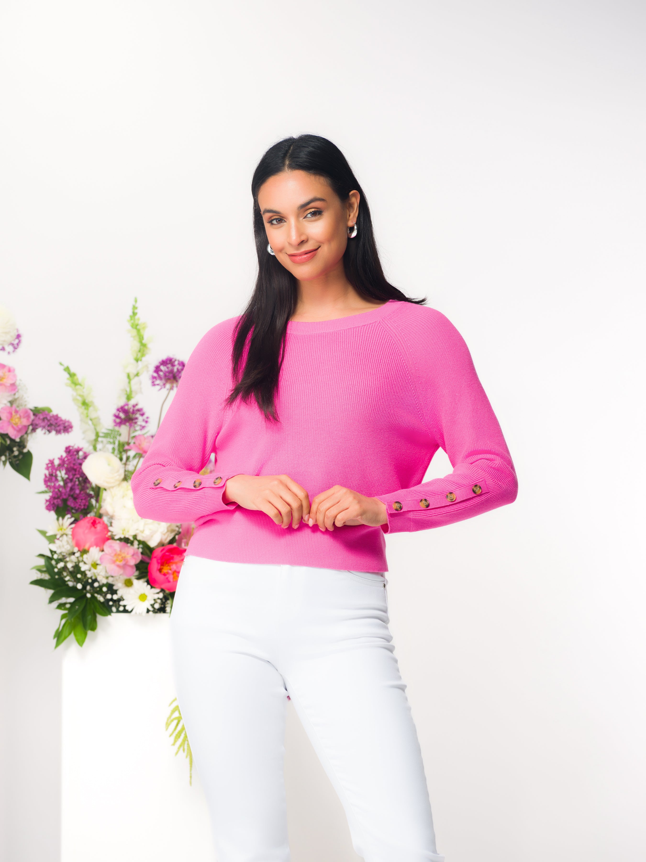Woman wearing a pink sweater with white pants standing next to a floral arrangement with a white background
