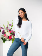 Woman wearing a white sweater with jeans standing next to a floral arrangement with a white background