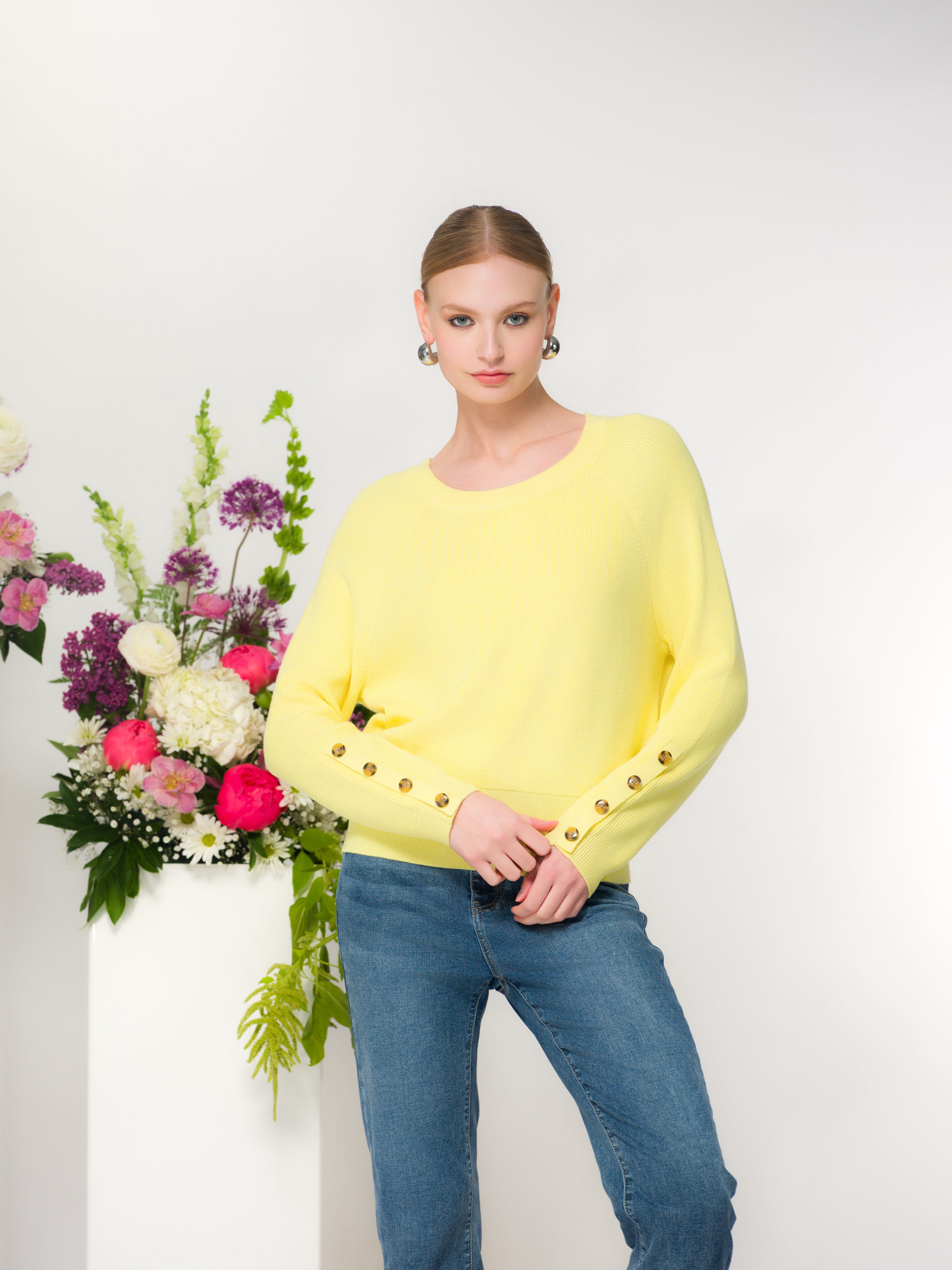 Woman wearing a yellow sweater with jeans standing next to a floral arrangement with a white background