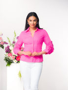 Woman with dark hair wearing a fuchsia blouse with white pants standing in front of a floral arrangement with a white background