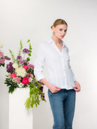Woman with blonde hair wearing a white blouse with denim pants standing in front of a floral arrangement with a white background