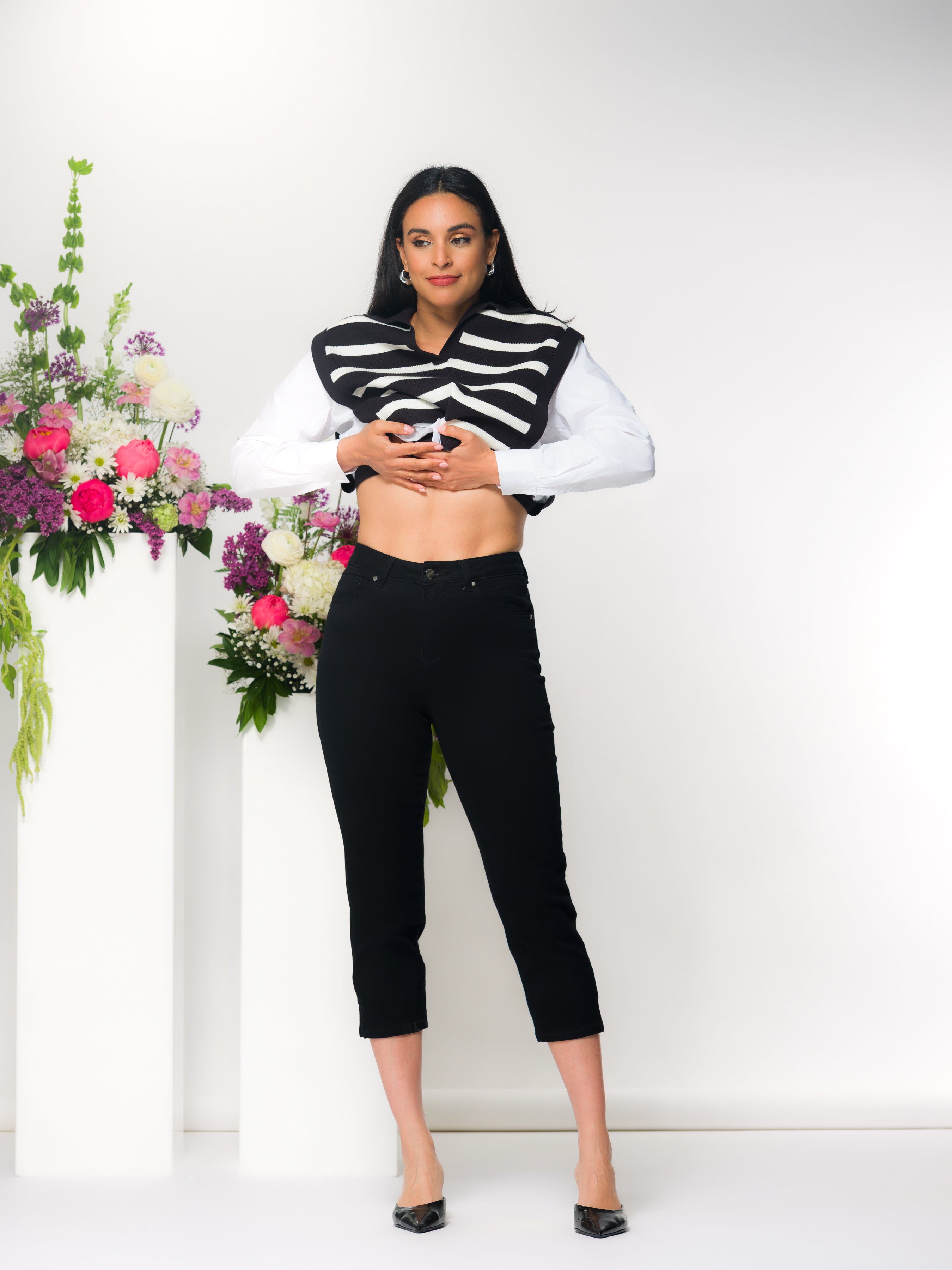 Dark haired woman wearing a sweater paired with black slim capri-length pants in a studio setting with floral arrangements on white pedestals.