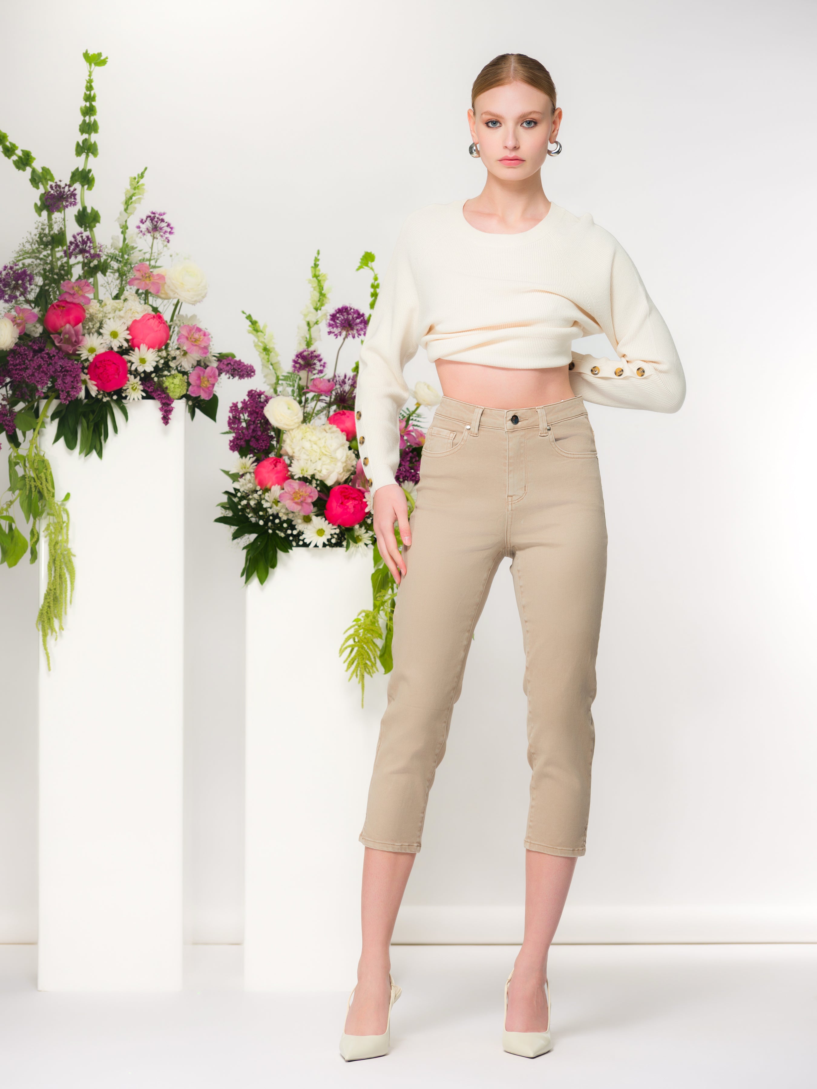 Light haired woman wearing a sweater paired with sand slim capri-length pants in a studio setting with floral arrangements on white pedestals.