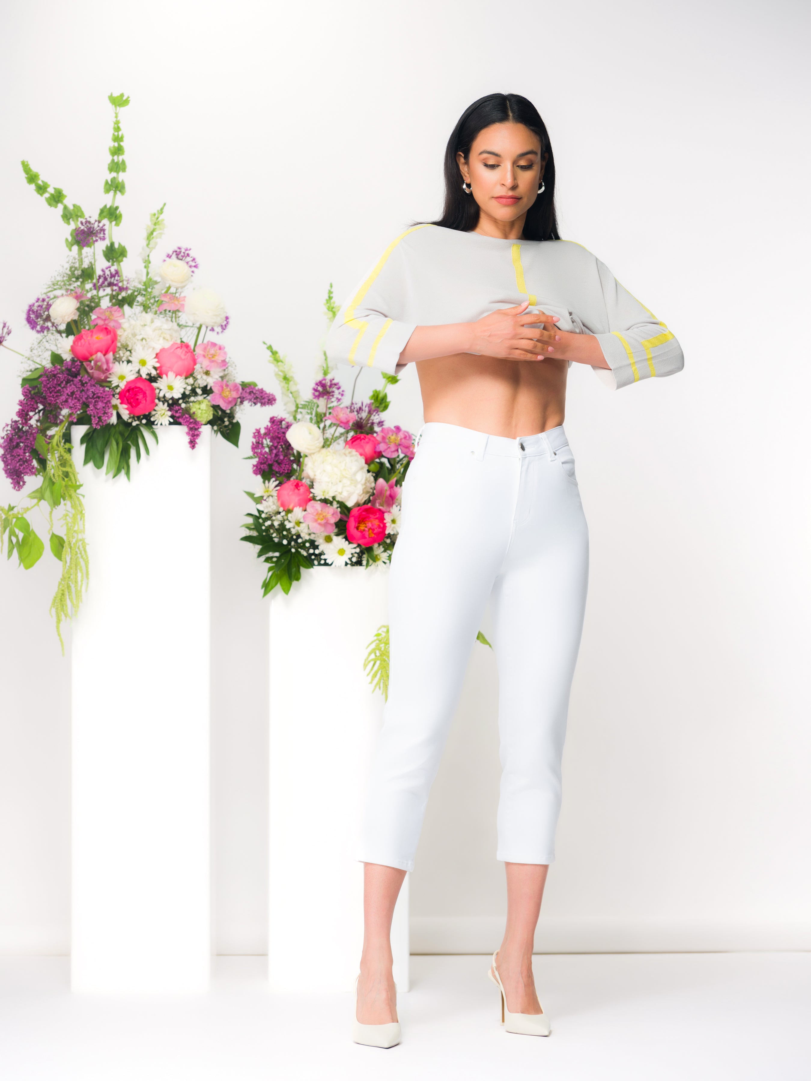 Dark haired woman wearing a sweater paired with white slim capri-length pants in a studio setting with floral arrangements on white pedestals.