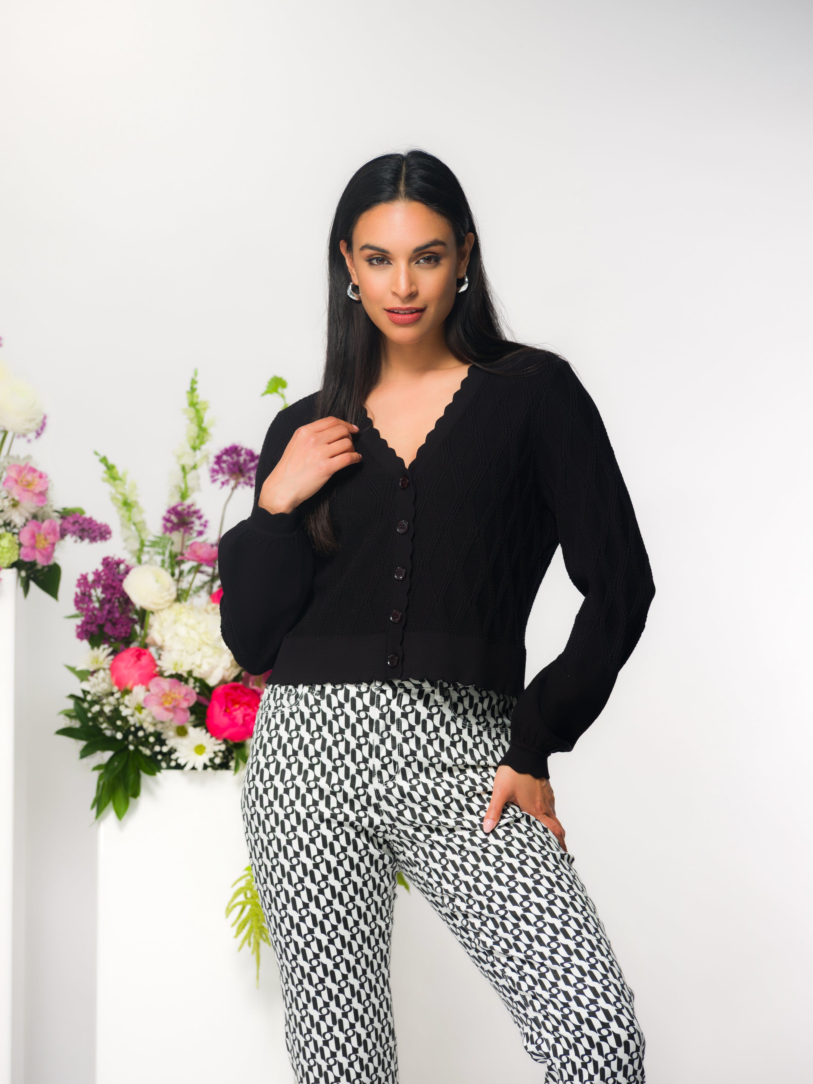 Model wearing a black fitted button-front knit cardigan with scalloped neckline and long sleeves, styled with pants, photographed in a studio setting.