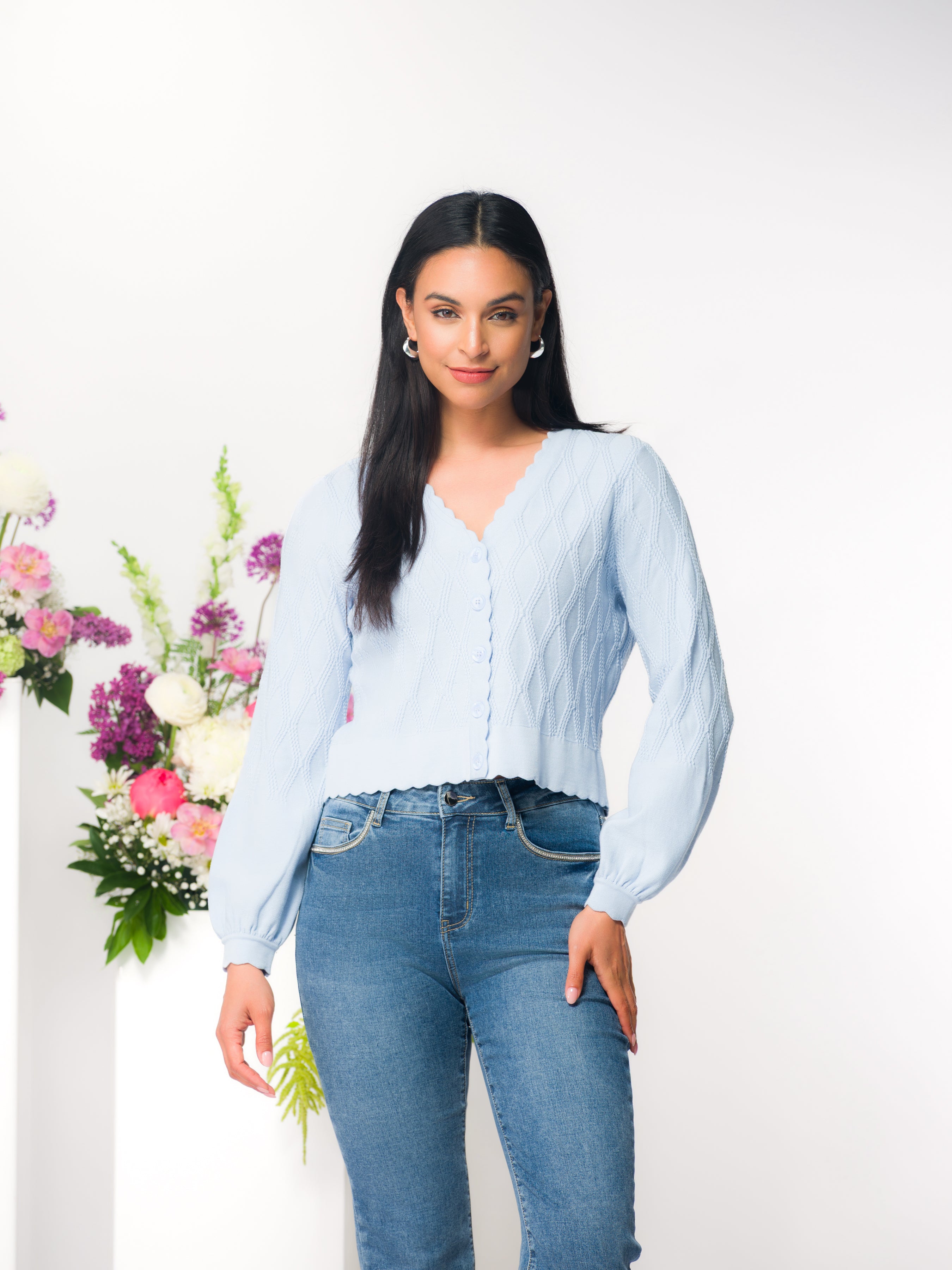 Model wearing a blue fitted button-front knit cardigan with scalloped neckline and long sleeves, styled with denim jeans, photographed in a studio setting.