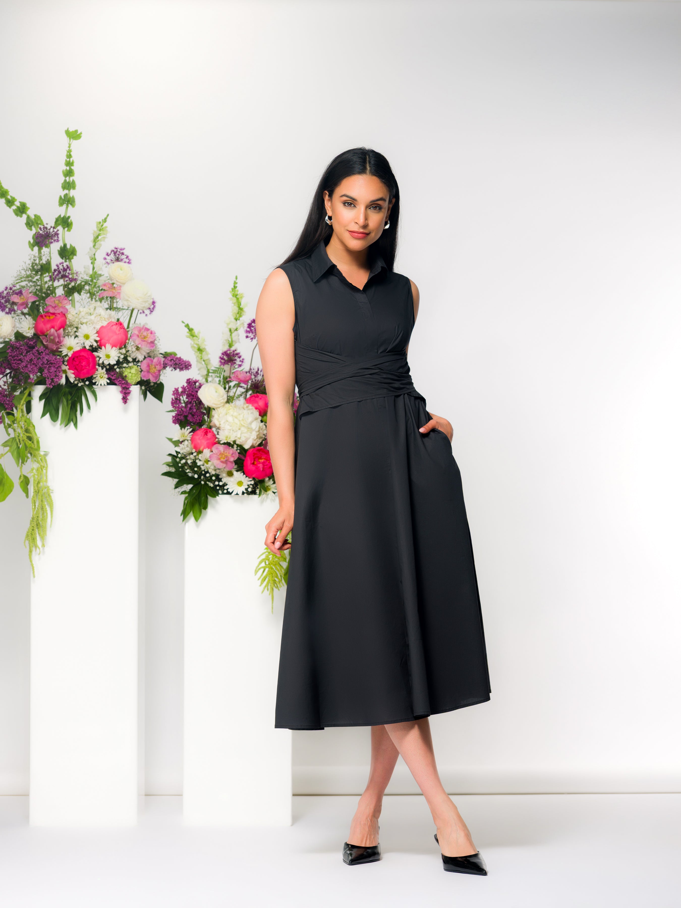 Model wearing a black sleeveless midi dress with a wrap-style waist and collared neckline, featuring a softly draped skirt and side pockets, styled with classic heels in a studio setting with floral accents.
