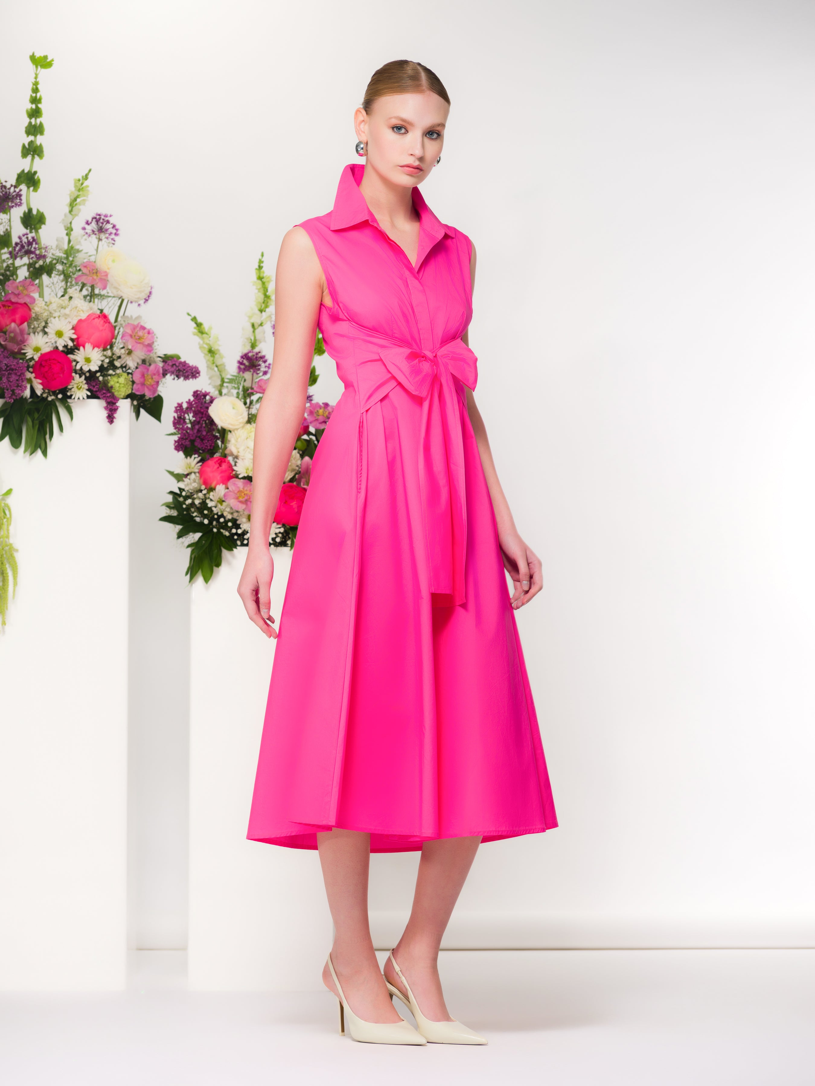 Model wearing a pink sleeveless midi dress with a wrap-style waist and collared neckline, featuring a softly draped skirt and side pockets, styled with classic heels in a studio setting with floral accents.