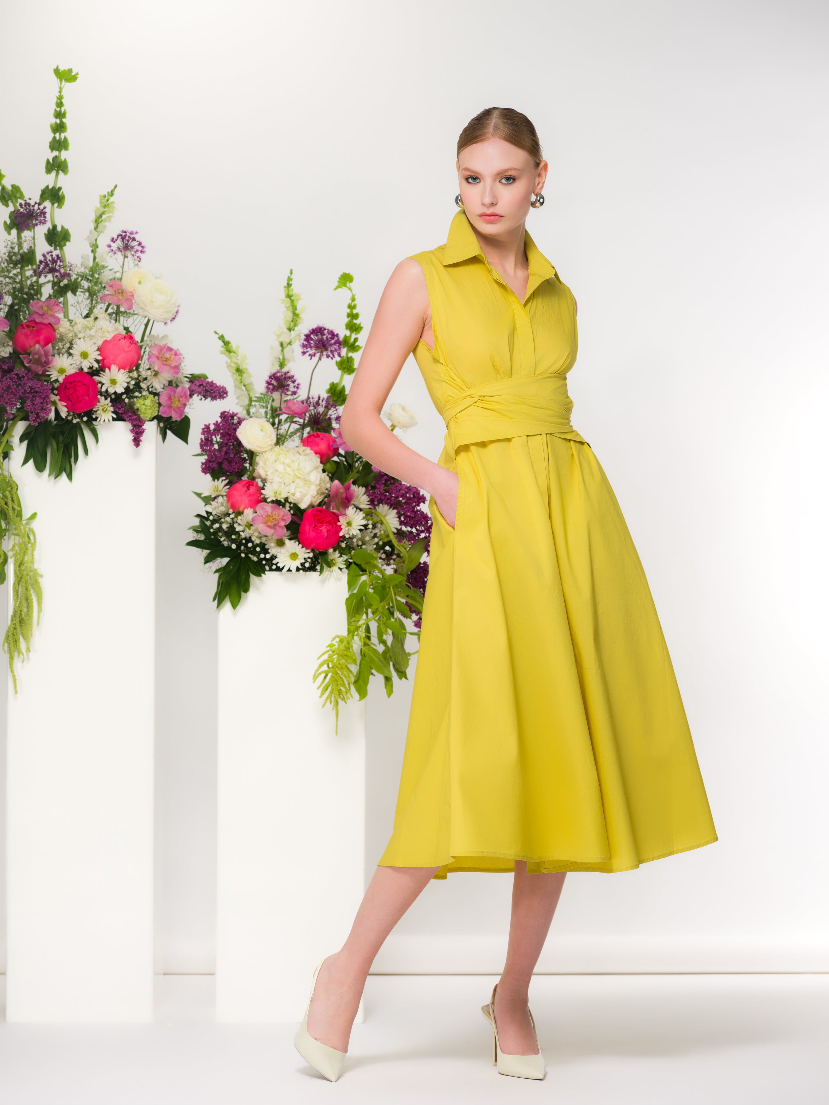 Model wearing a lime greensleeveless midi dress with a wrap-style waist and collared neckline, featuring a softly draped skirt and side pockets, styled with classic heels in a studio setting with floral accents.