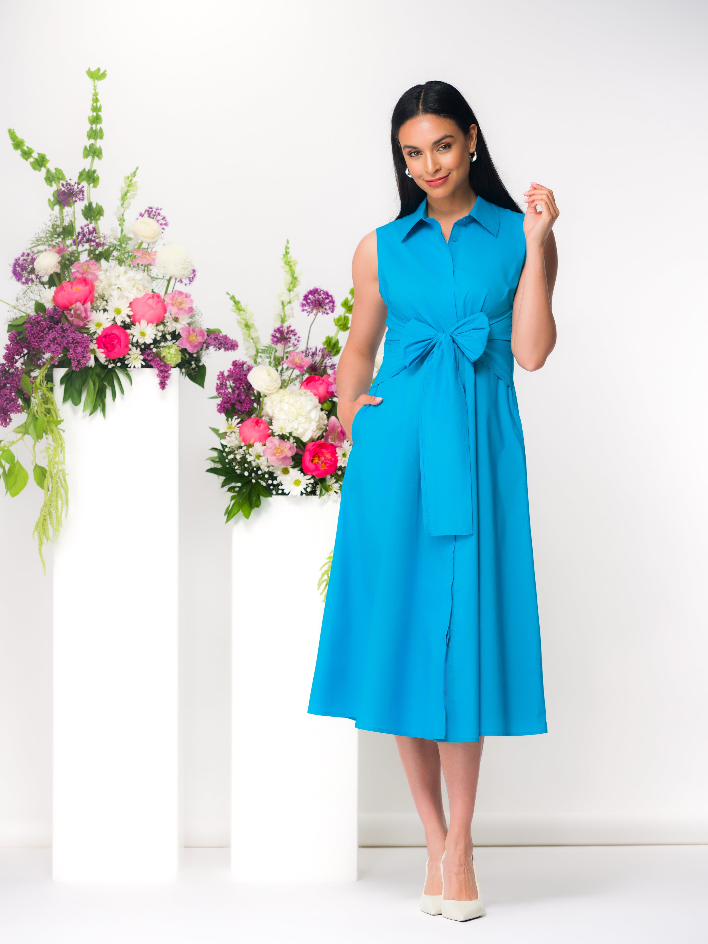 Model wearing a turquoise sleeveless midi dress with a wrap-style waist and collared neckline, featuring a softly draped skirt and side pockets, styled with classic heels in a studio setting with floral accents.