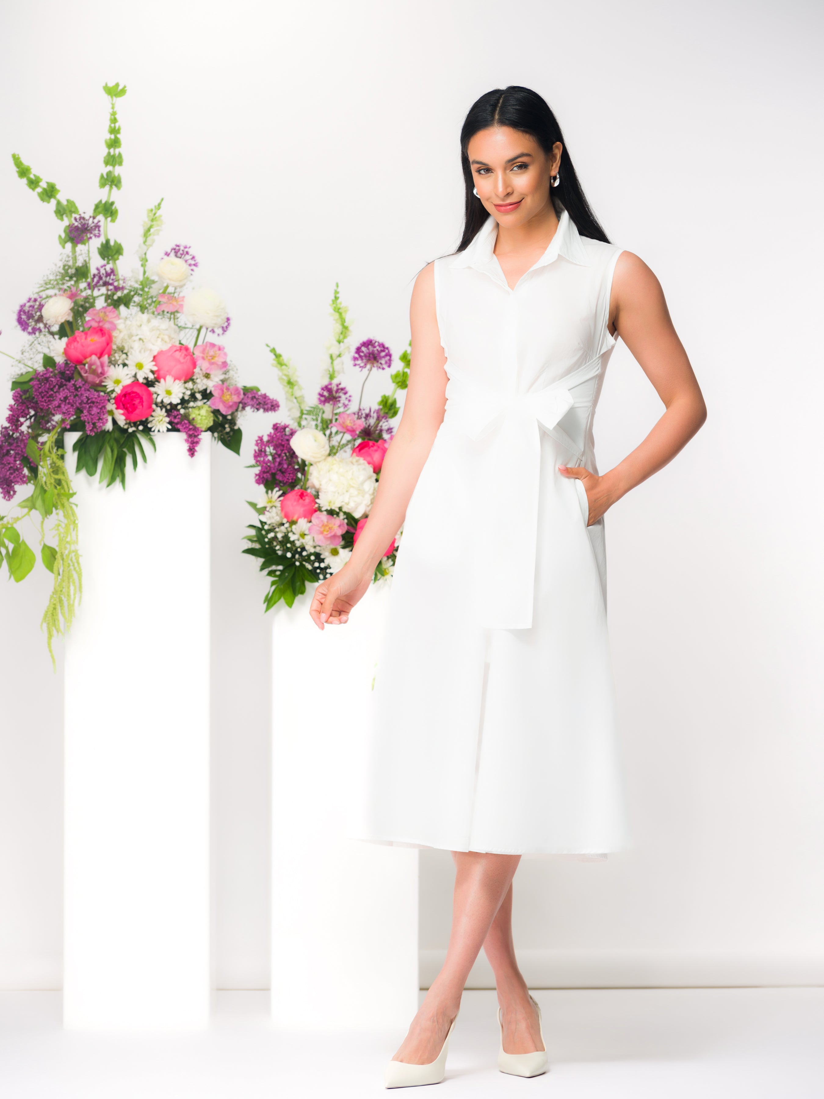 Model wearing a white sleeveless midi dress with a wrap-style waist and collared neckline, featuring a softly draped skirt and side pockets, styled with classic heels in a studio setting with floral accents.