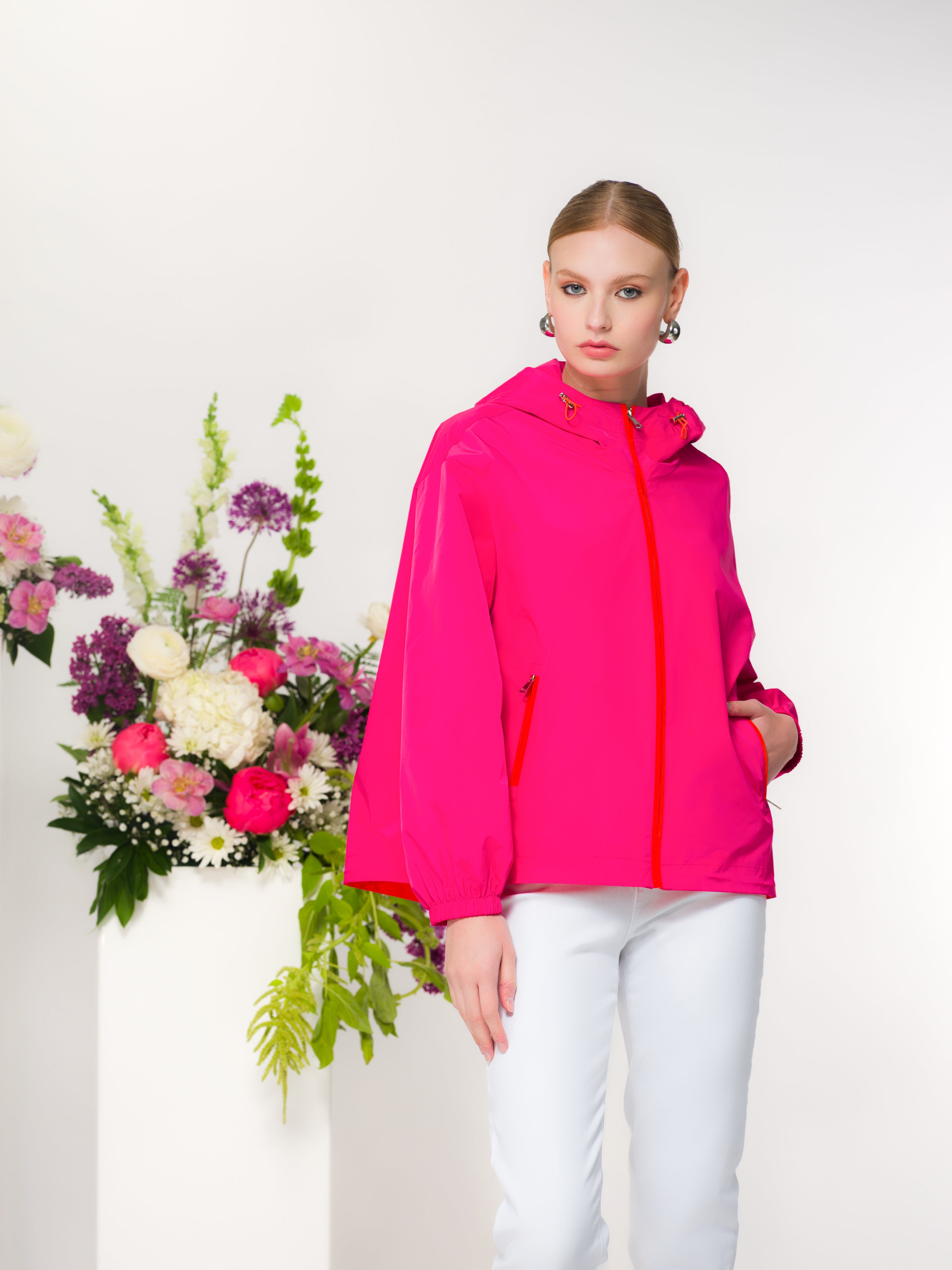 Woman wearing a bright pink jacket with white pants standing next to floral arrangements on a white background