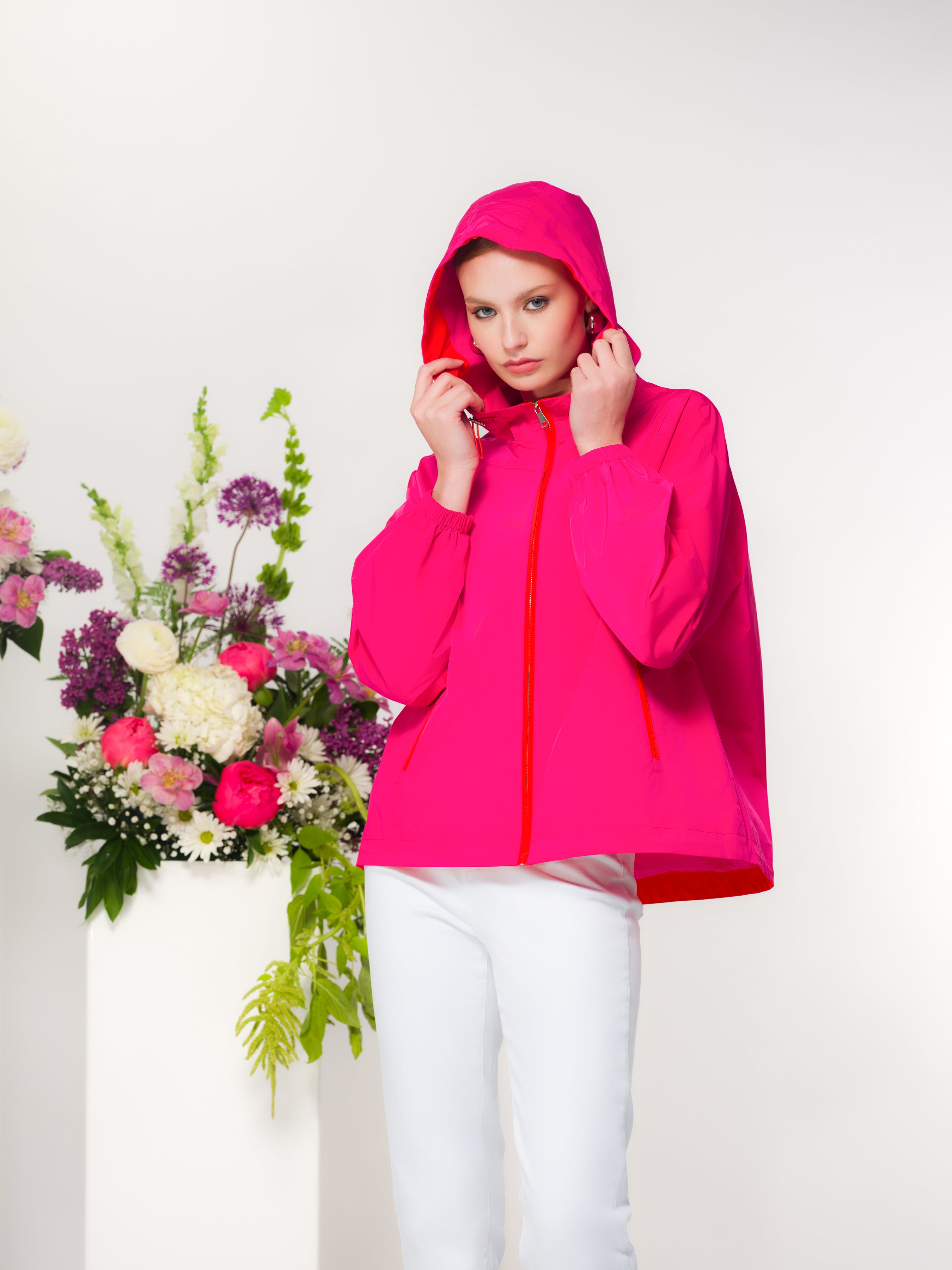 Woman wearing a bright pink jacket with white pants standing next to floral arrangements on a white background