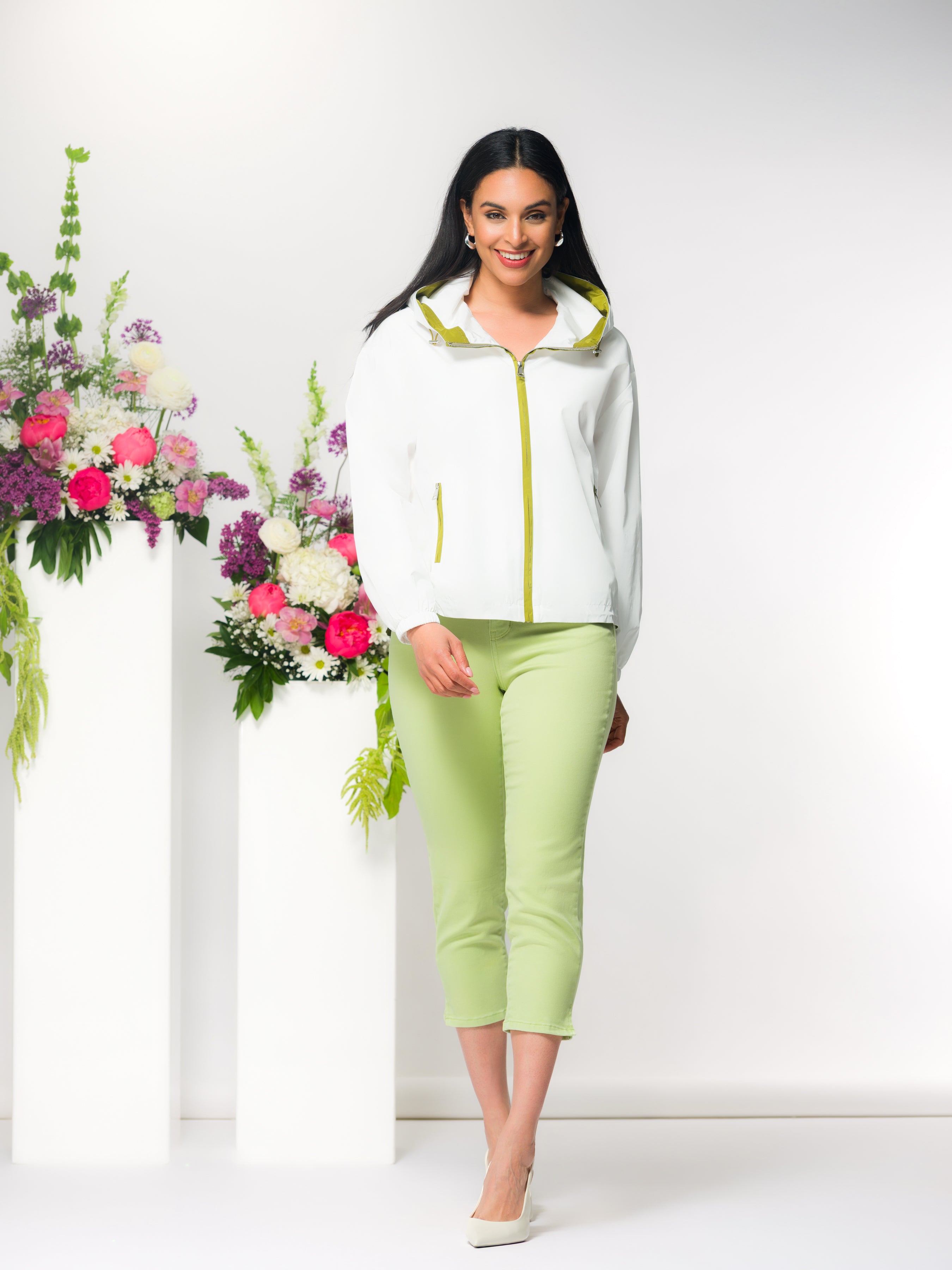 Woman wearing a white jacket with green cropped pants standing next to floral arrangements on a white background