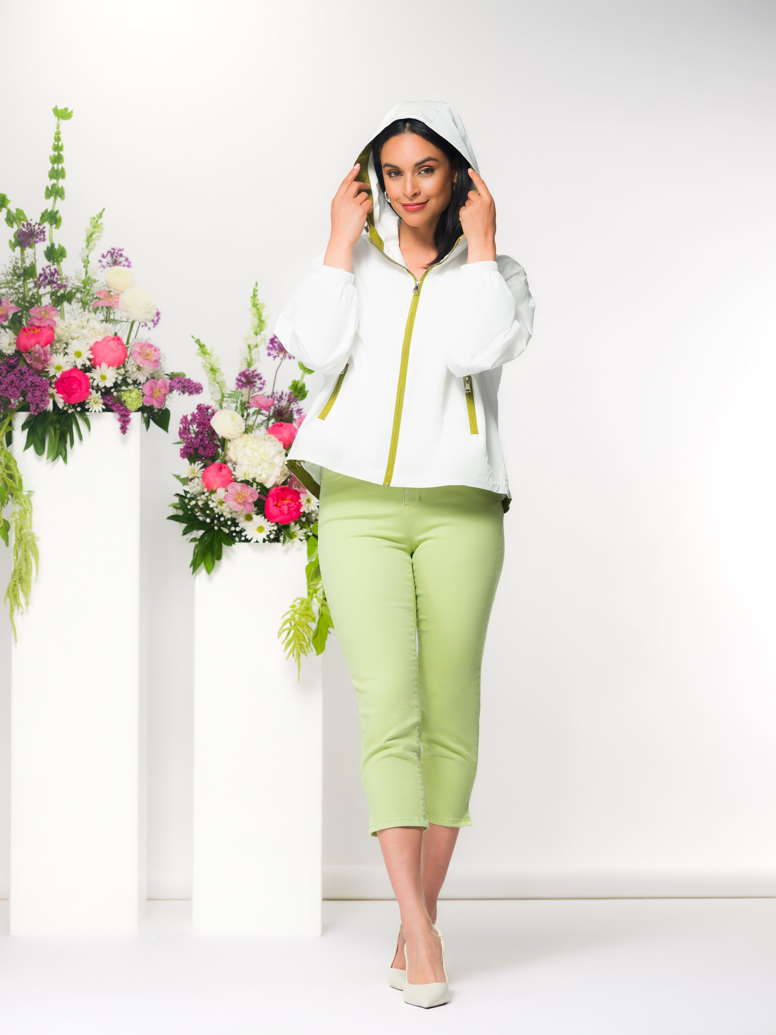 Woman wearing a white jacket with green cropped pants standing next to floral arrangements on a white background