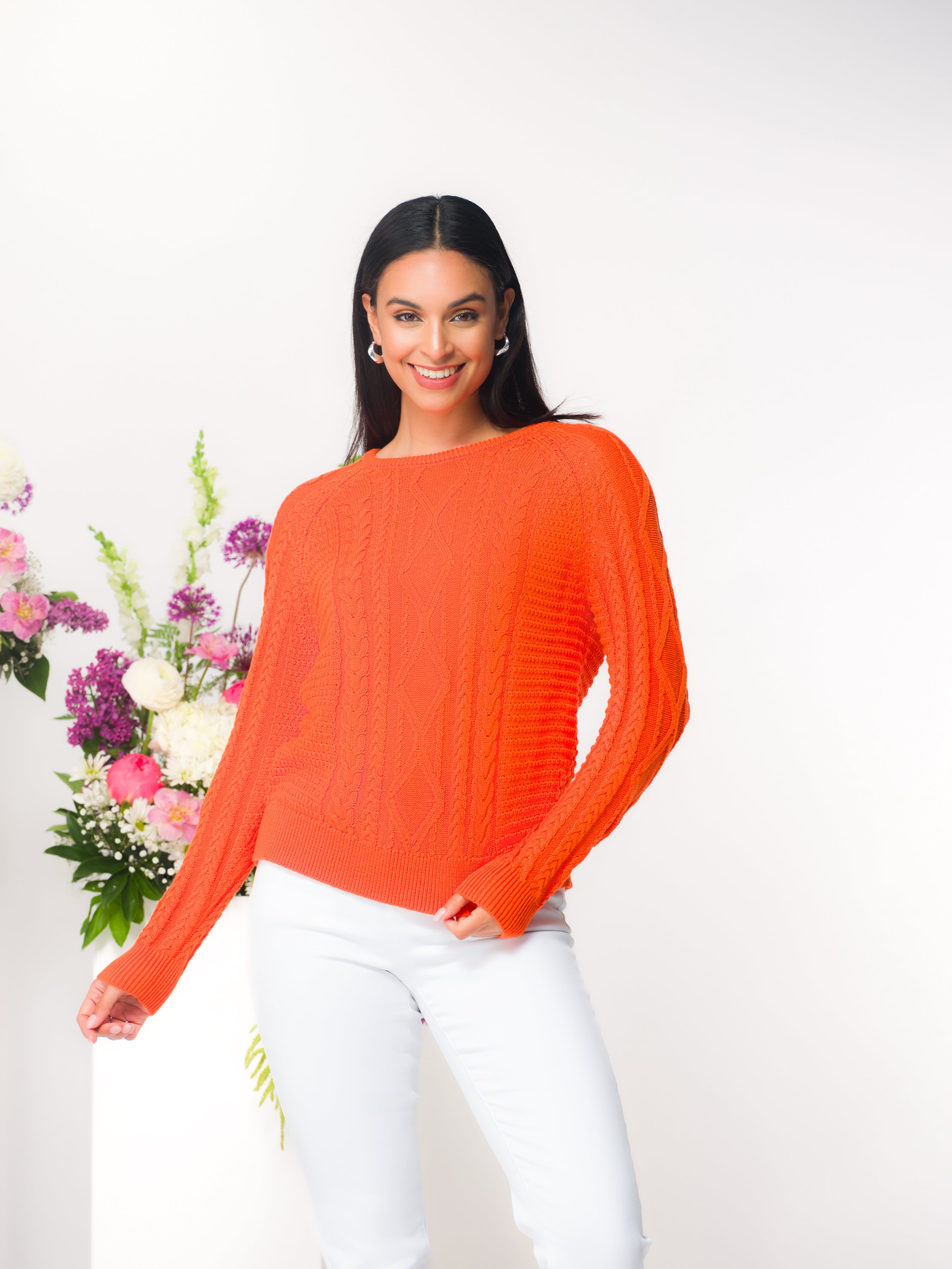 Model wearing an orange cable knit pullover sweater with a crew neckline and long sleeves, styled with white pants in a studio setting.