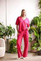 Woman wearing a relaxed pink V-neck top with short sleeves and matching wide-leg pants, styled indoors with plants.