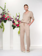 Woman wearing a relaxed sand coloured V-neck top with short sleeves and matching wide-leg pants, styled indoors with floral arrangements.